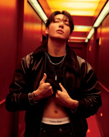 Adult man with lip piercing poses in a black leather jacket and jeans, lifting his black t-shirt to reveal underwear in a studio.