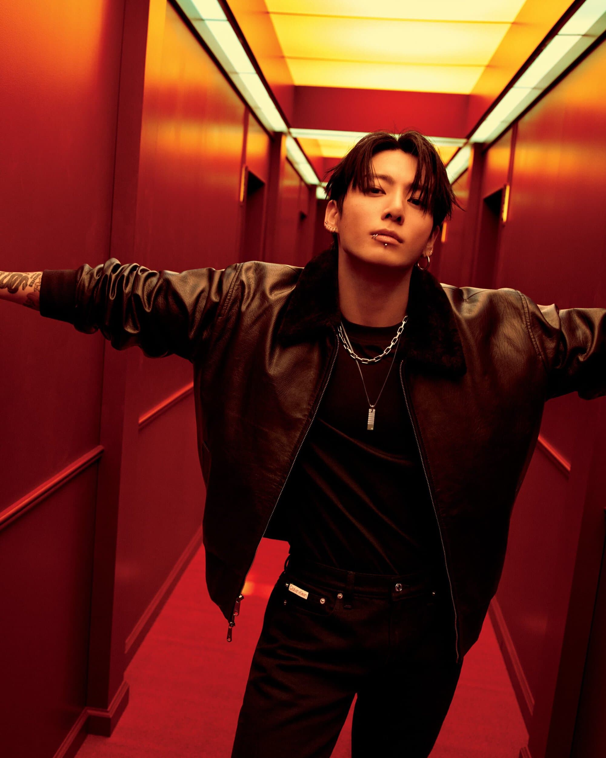 Stylish young man in a brown leather jacket and black shirt strikes a pose in a red hallway with yellow lighting, looking at the camera.