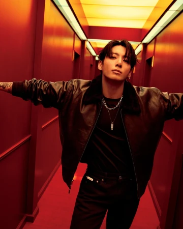 Stylish young man in a brown leather jacket and black shirt strikes a pose in a red hallway with yellow lighting, looking at the camera.