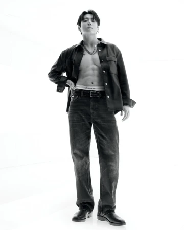 Black and white portrait of a muscular male fashion model standing indoors wearing jeans and an open jacket, against a stark white backdrop.