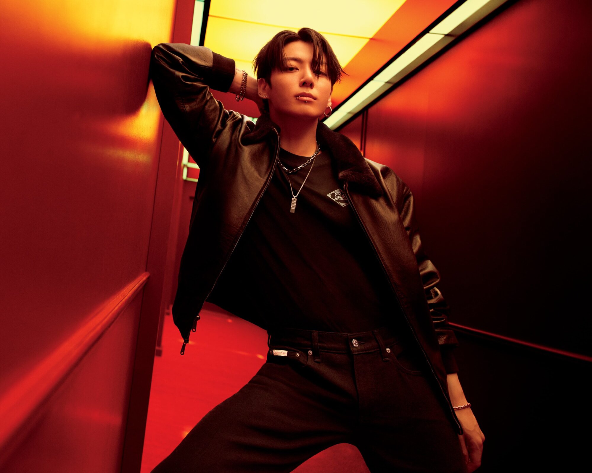 Portrait of a young adult man in a black leather jacket and jeans posing in a hallway with red and orange walls and ceiling lights.