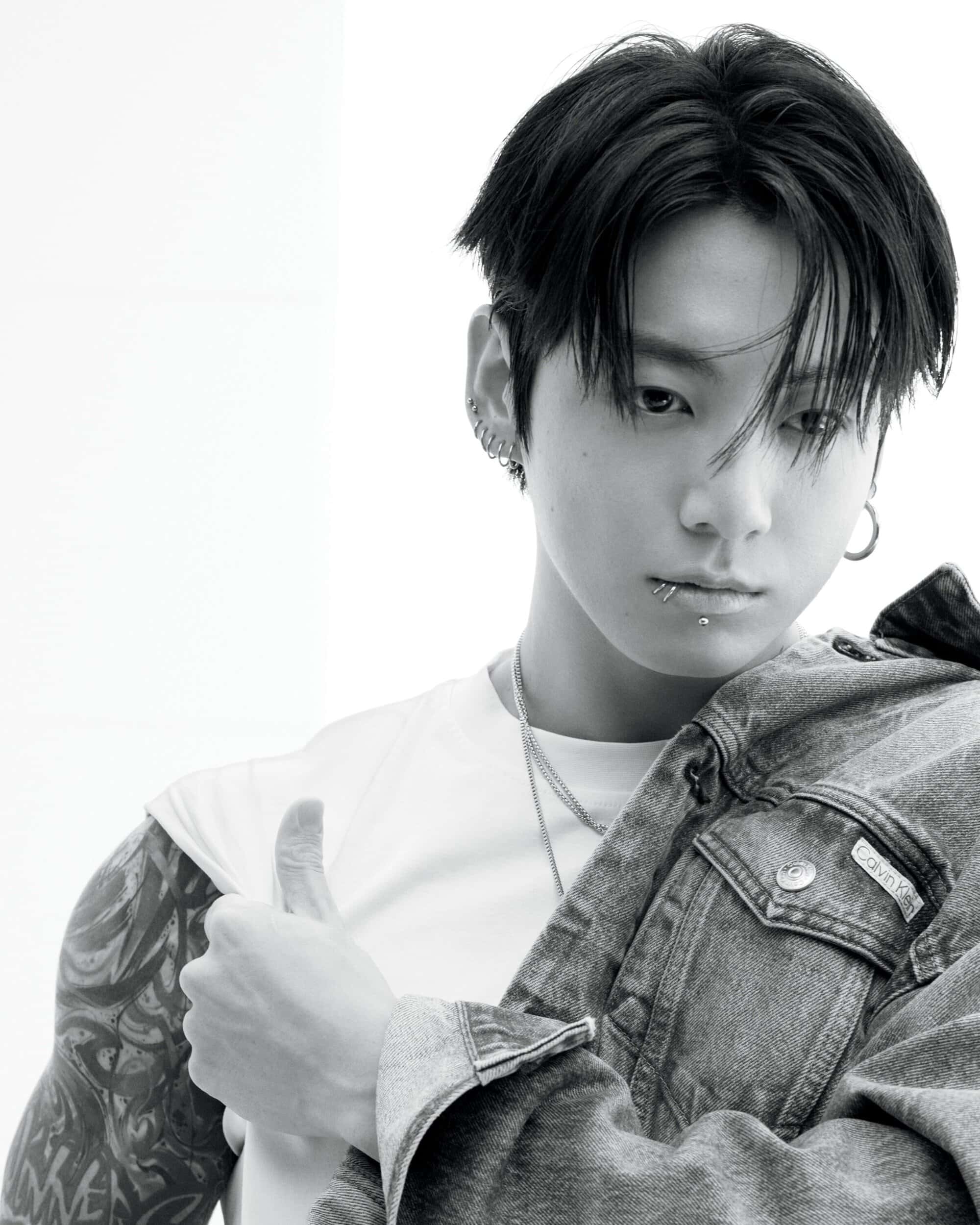 Monochrome close-up portrait of a young adult man with lip piercings, tattoos, and a denim jacket, posing against a bright white background.