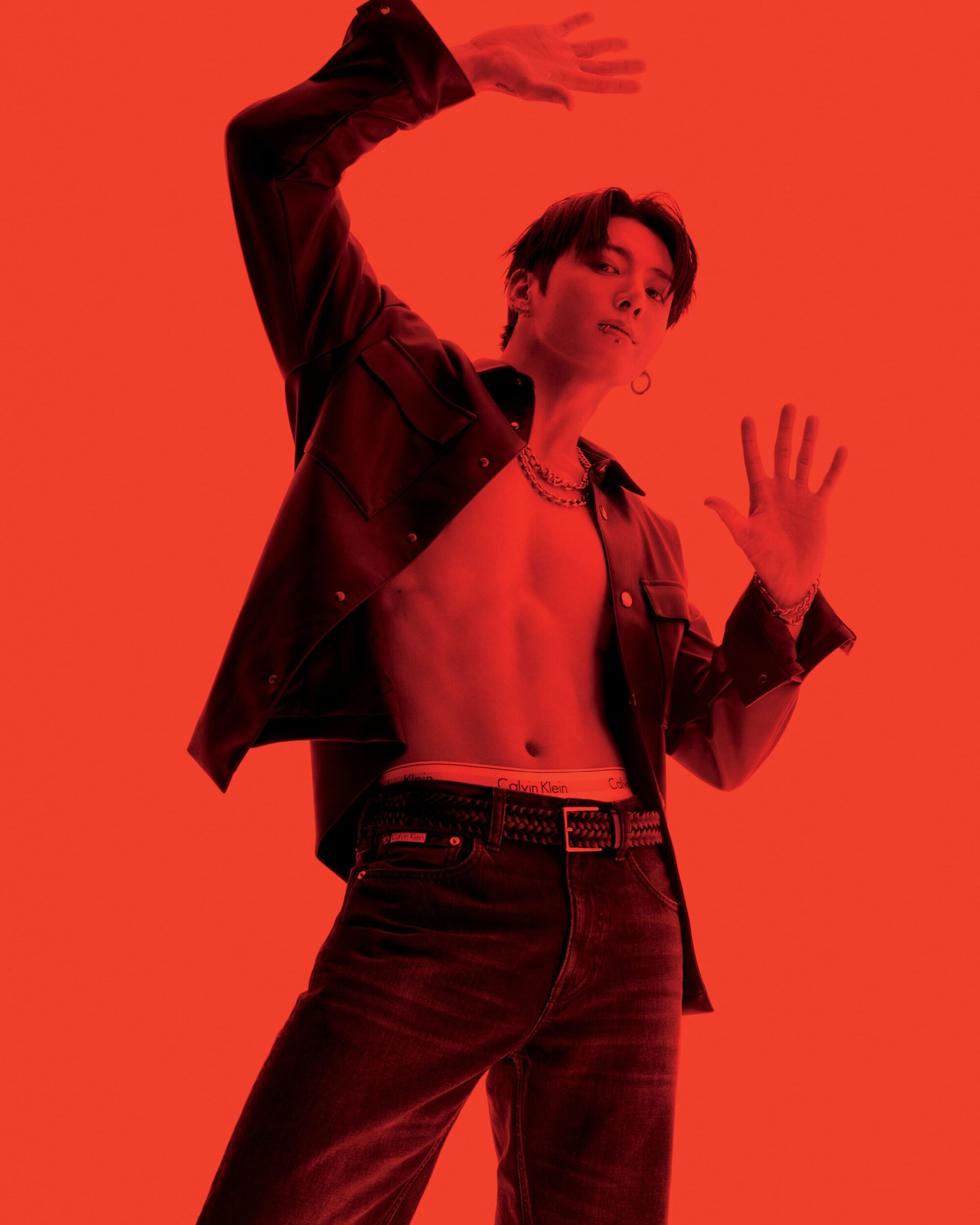 An adult male in a red-toned studio, shirtless with an open jacket, jeans, and a belt, posing with arms raised in a low-angle full shot.