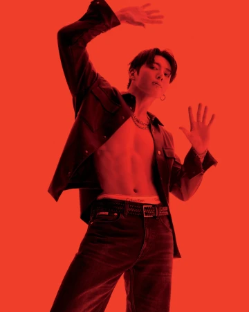 An adult male in a red-toned studio, shirtless with an open jacket, jeans, and a belt, posing with arms raised in a low-angle full shot.