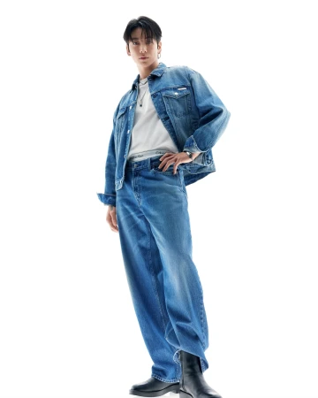 Jung Kook wearing a blue denim jacket and matching jeans against a bright white background for Calvin Klein.