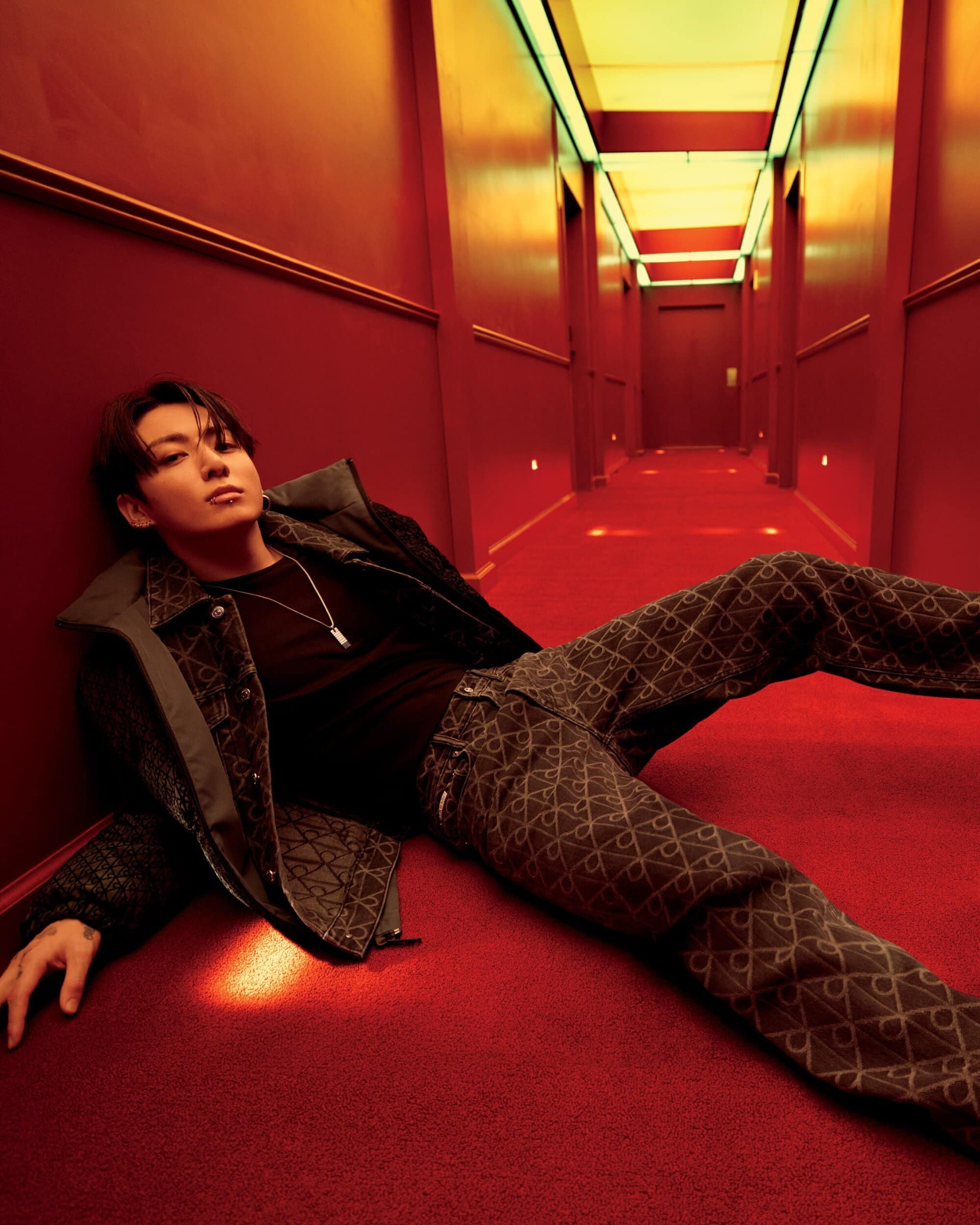 A young man in a dark patterned outfit lies on a red carpet in a long red hallway.