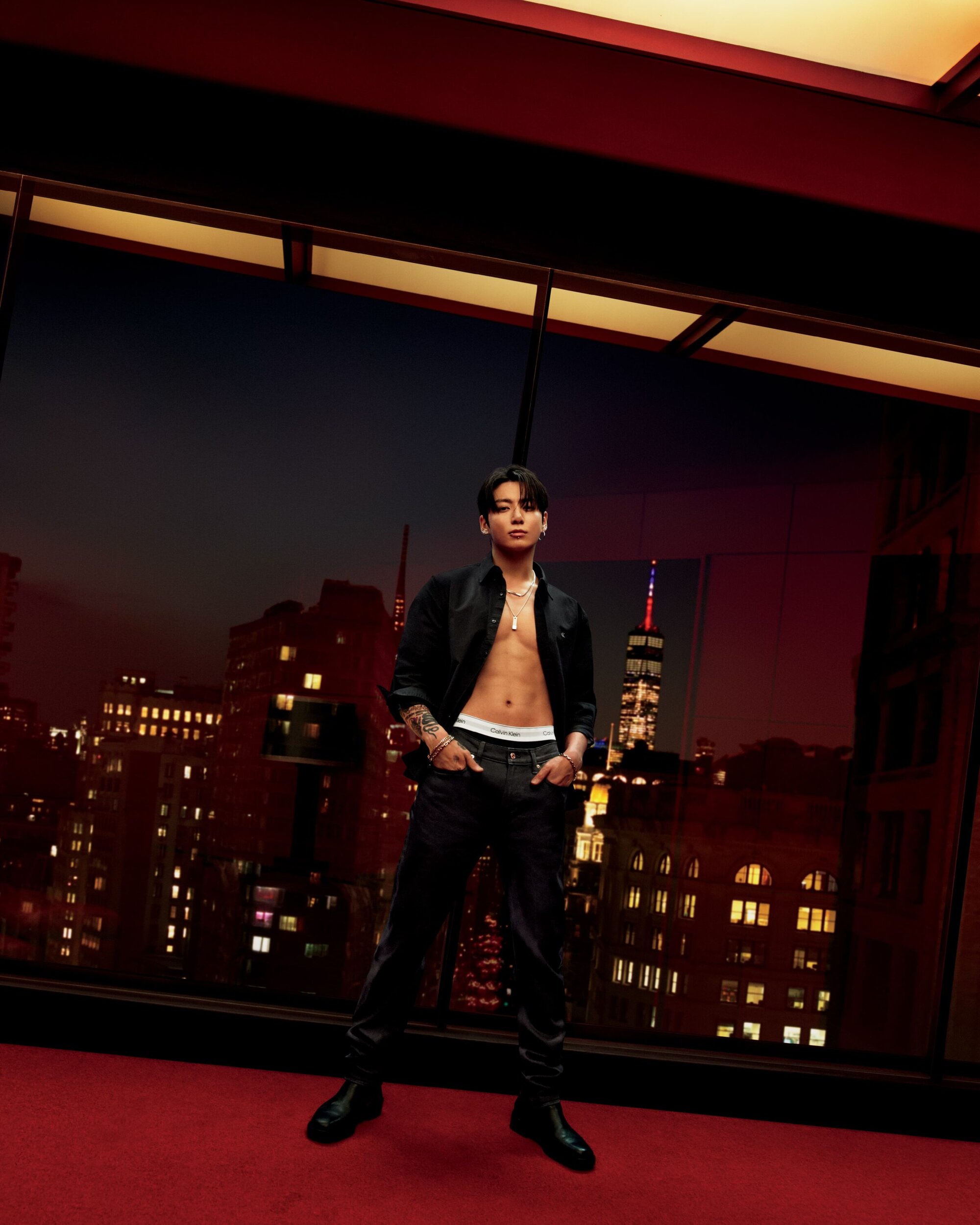 Male model in black jacket and pants standing against a window overlooking a cityscape at dusk, fashion-forward pose and style.
