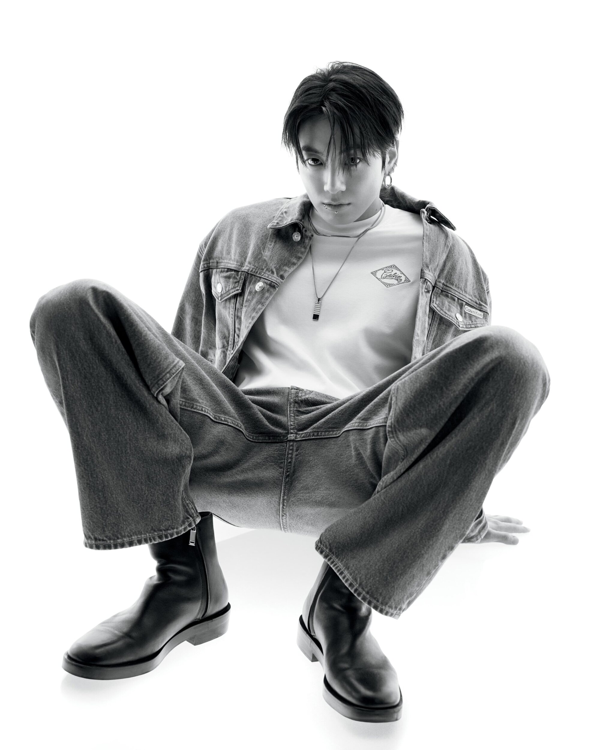 Jung Kook in a denim jacket and wide-leg jeans squatting against a white background for Calvin Klein.