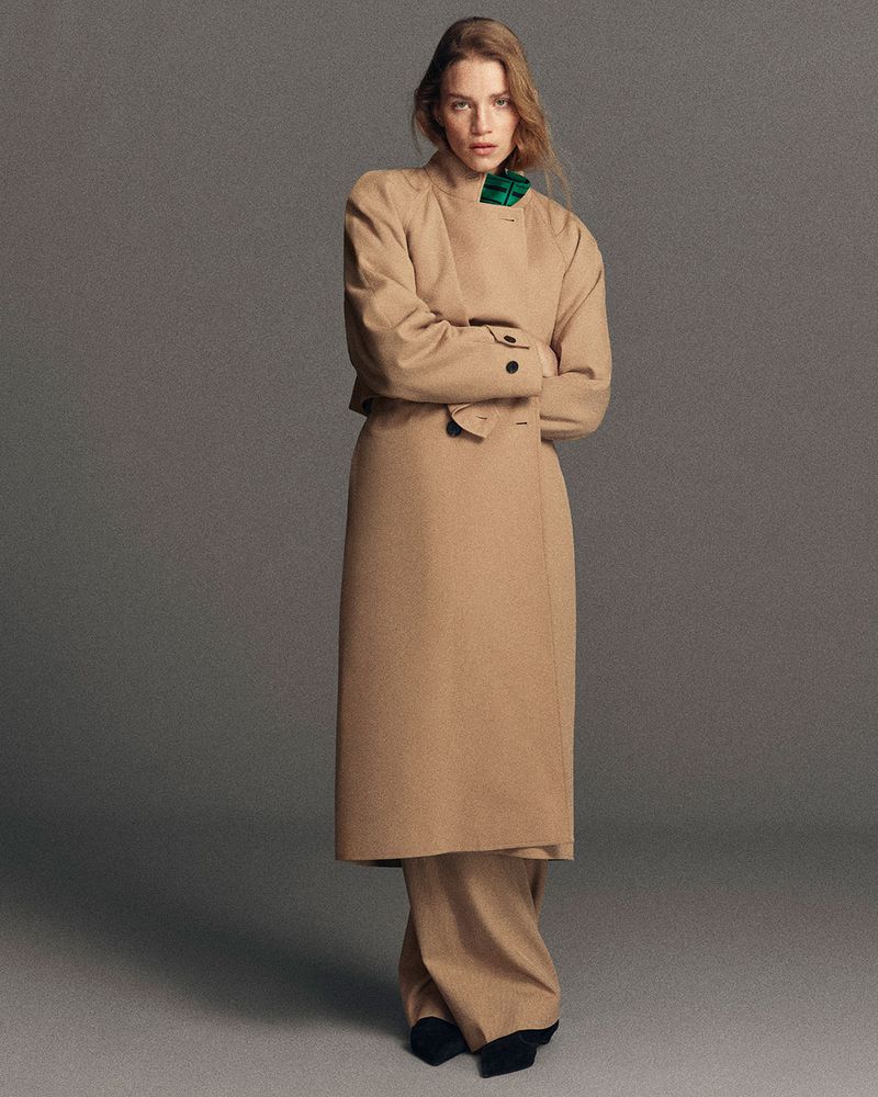 Full-length fashion shot of a woman in a tan trench coat and wide-leg pants, standing with arms crossed in a studio setting, neutral backdrop.