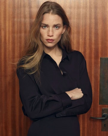 Portrait of an adult woman with long blonde hair and a serious expression, wearing a black collared shirt and standing indoors.