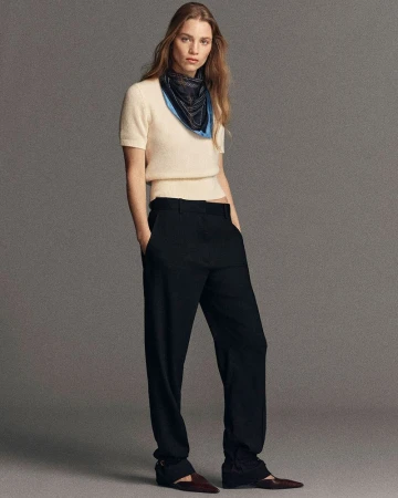 Full-length portrait of an adult female fashion model in black trousers, off-white sweater, brown shoes, and scarf against a neutral backdrop.