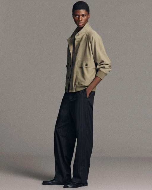Male fashion model poses in khaki bomber jacket and pinstripe trousers against neutral gray background in a studio setting.