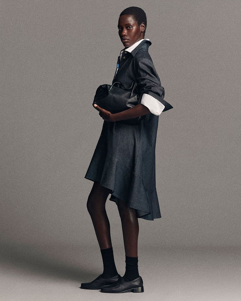 Model in dark denim dress holding black leather bag against a neutral grey studio background