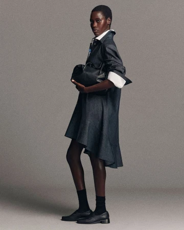 Model in dark denim dress holding black leather bag against a neutral grey studio background