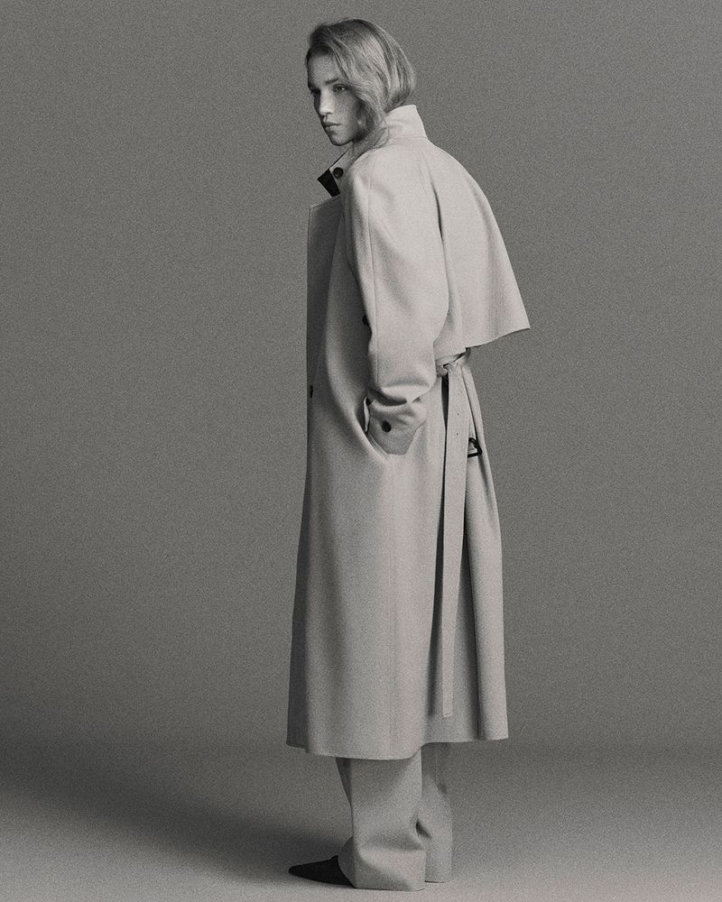 Full-length shot of an adult female with fair skin and blonde hair wearing a neutral trench coat and standing in a photo studio.