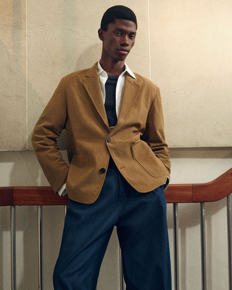 A male model wearing a camel blazer and dark blue trousers leaning against a wall