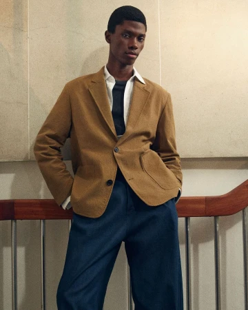 A male model wearing a camel blazer and dark blue trousers leaning against a wall