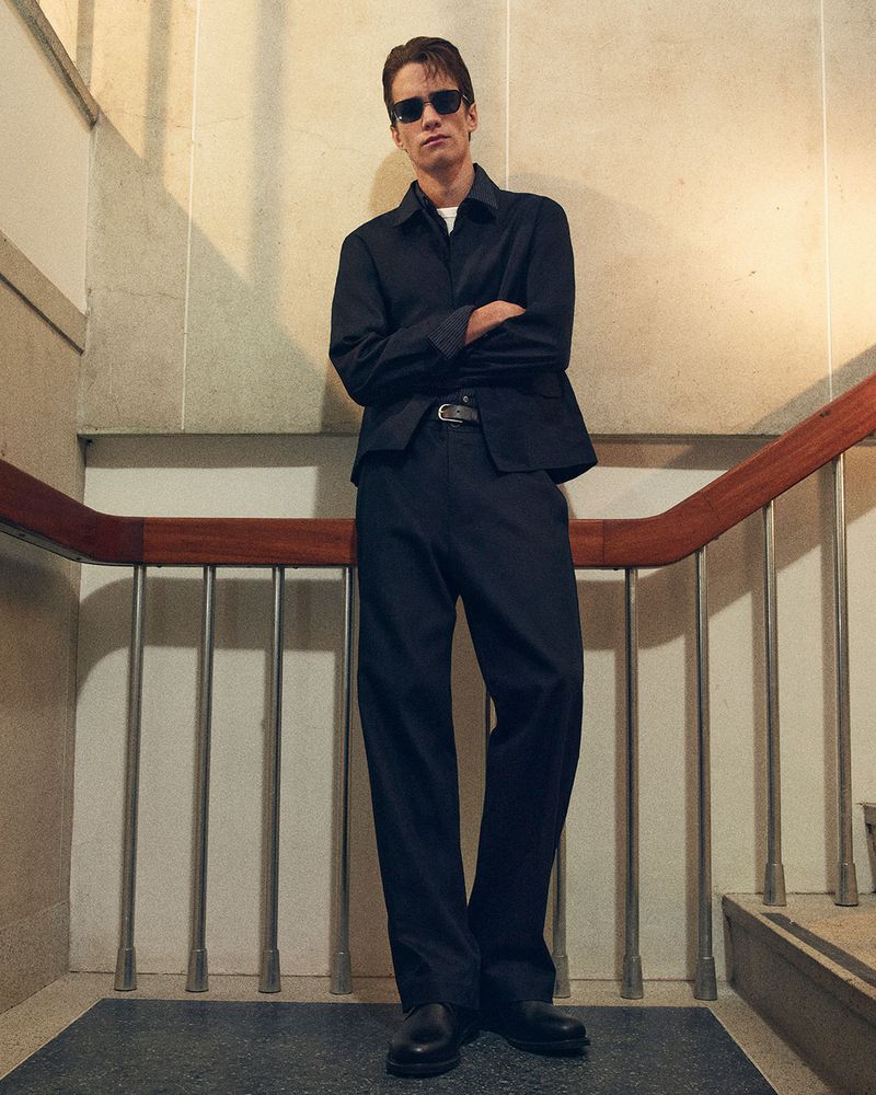 A young adult man in a dark blue jacket and pants stands on a staircase, complemented by light-colored walls and natural lighting.
