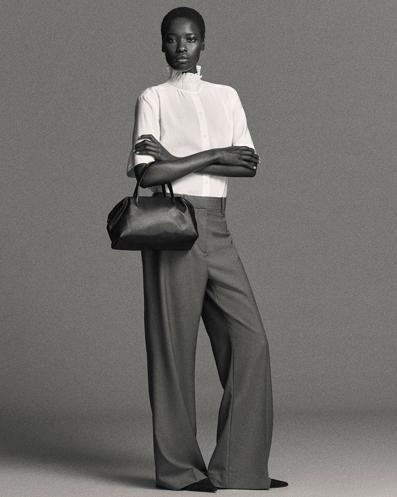 Elegant Black woman poses in a white blouse, gray trousers, and black purse against a gray studio backdrop in a full studio shot.
