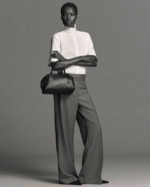 Elegant Black woman poses in a white blouse, gray trousers, and black purse against a gray studio backdrop in a full studio shot.