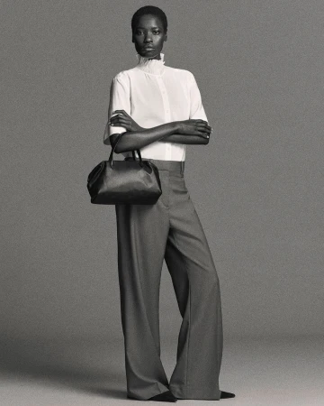 Elegant Black woman poses in a white blouse, gray trousers, and black purse against a gray studio backdrop in a full studio shot.