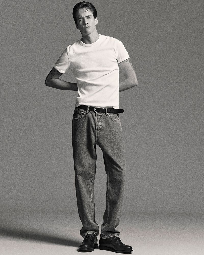 Studio shot of a young adult male in a white tee and jeans, standing with arms behind his back, minimalist aesthetic on a grey backdrop.