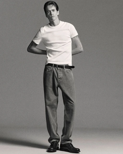 Studio shot of a young adult male in a white tee and jeans, standing with arms behind his back, minimalist aesthetic on a grey backdrop.