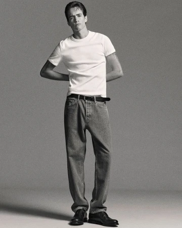 Studio shot of a young adult male in a white tee and jeans, standing with arms behind his back, minimalist aesthetic on a grey backdrop.