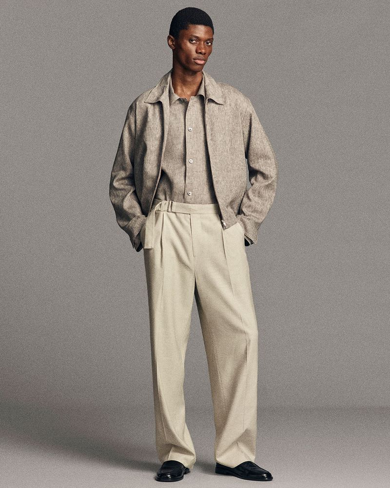 Full-length shot of an African-American male model wearing a light brown jacket, button-down shirt, beige pleated pants and black loafers.