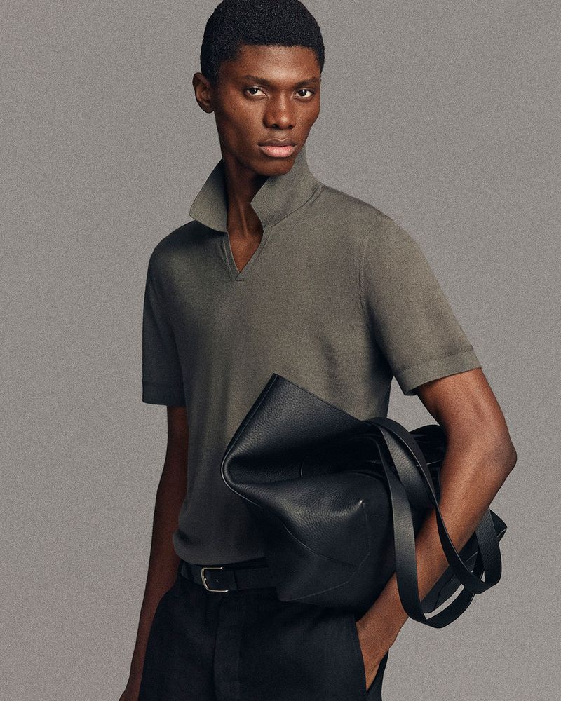 Fashionable African-American man in polo shirt holds black leather bag, wearing black pants and belt, posing against neutral backdrop.