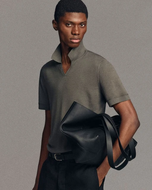 Fashionable African-American man in polo shirt holds black leather bag, wearing black pants and belt, posing against neutral backdrop.