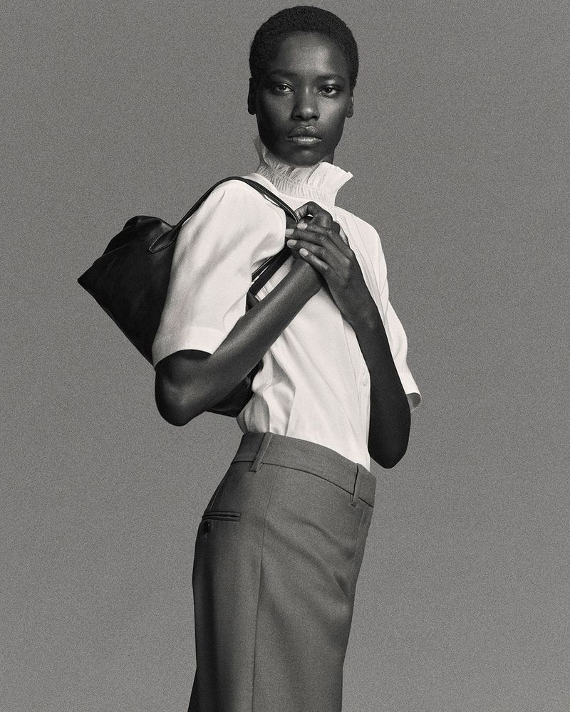 An adult woman poses in a studio wearing a white short-sleeve shirt, grey skirt, and black shoulder bag against a grey backdrop.