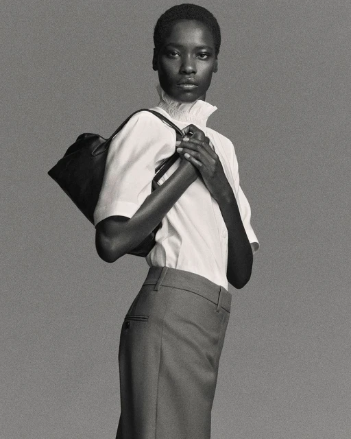 An adult woman poses in a studio wearing a white short-sleeve shirt, grey skirt, and black shoulder bag against a grey backdrop.