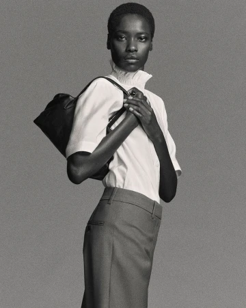 An adult woman poses in a studio wearing a white short-sleeve shirt, grey skirt, and black shoulder bag against a grey backdrop.