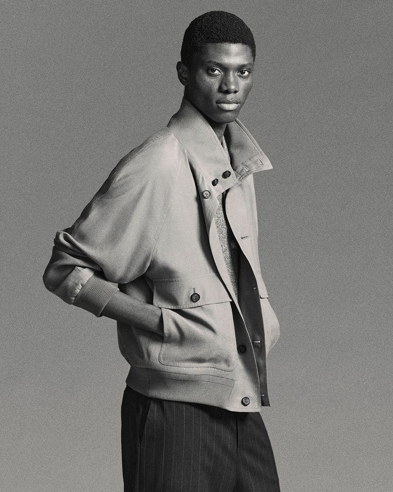 Portrait of an African American adult male wearing a light brown jacket and dark striped pants, captured in a studio setting.