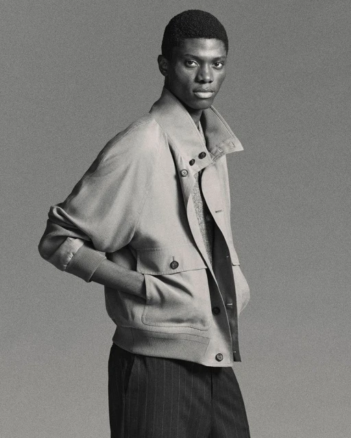 Portrait of an African American adult male wearing a light brown jacket and dark striped pants, captured in a studio setting.