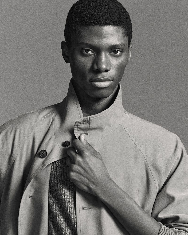 Monochrome portrait of an African American man wearing a collared jacket, standing indoors against a solid grey backdrop. Beauty and fashion.
