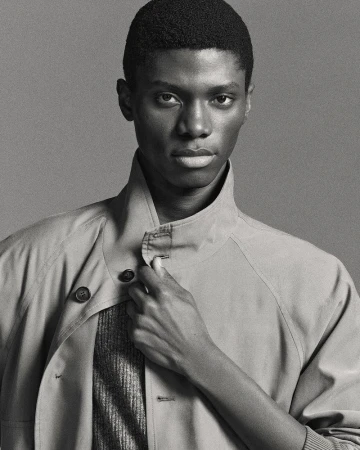 Monochrome portrait of an African American man wearing a collared jacket, standing indoors against a solid grey backdrop. Beauty and fashion.