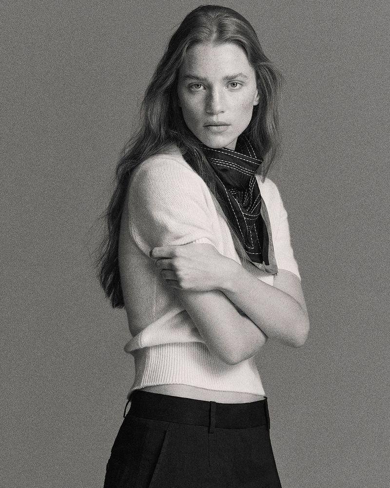 Black and white portrait of a woman in a short-sleeved sweater and black pants with a scarf around her neck in a studio setting.