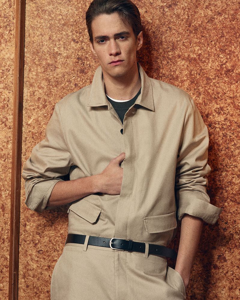 Portrait of a young adult male with brown hair wearing a khaki jumpsuit and a black belt, posed against a textured brown wall.
