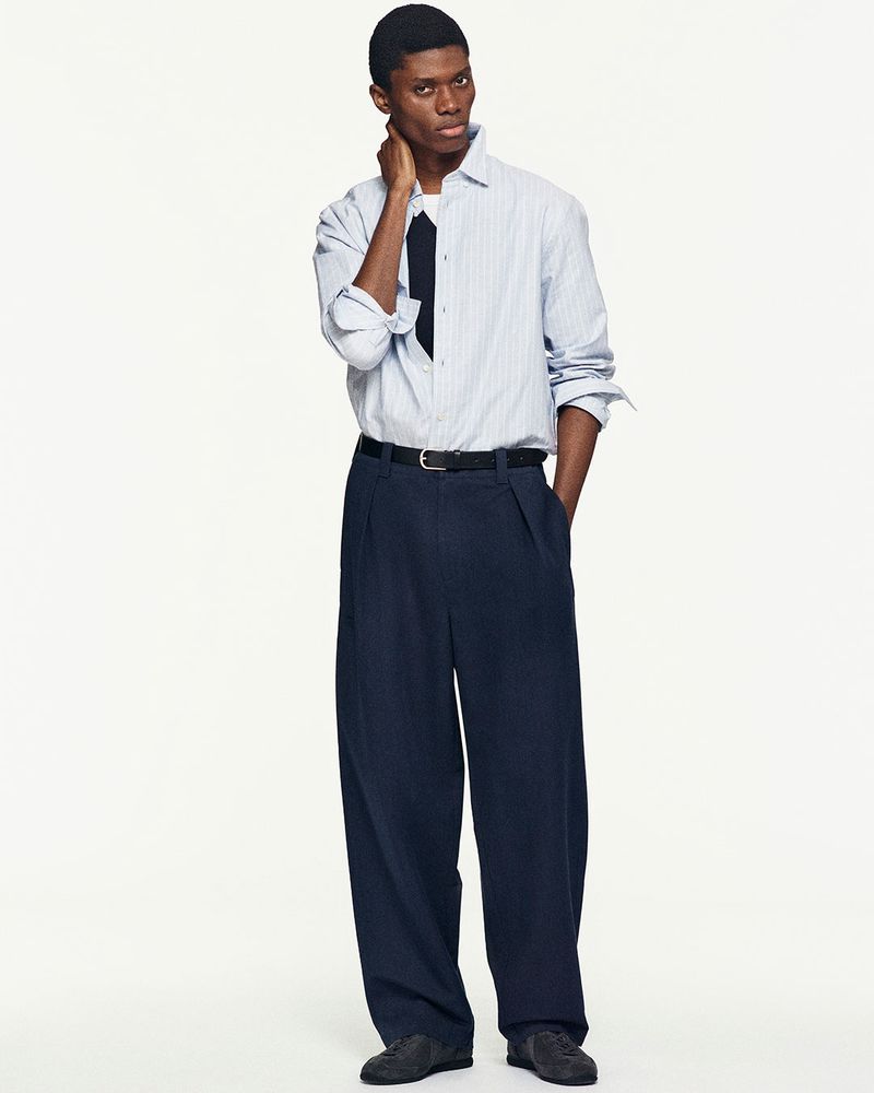 Full-length shot of a Black male model in a light-blue striped shirt, navy tie, and oversized navy pants against a white backdrop.