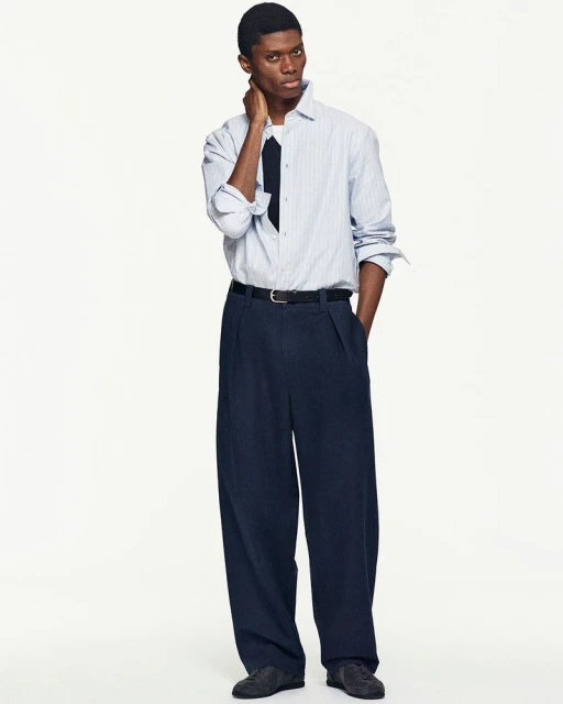 Full-length shot of a Black male model in a light-blue striped shirt, navy tie, and oversized navy pants against a white backdrop.