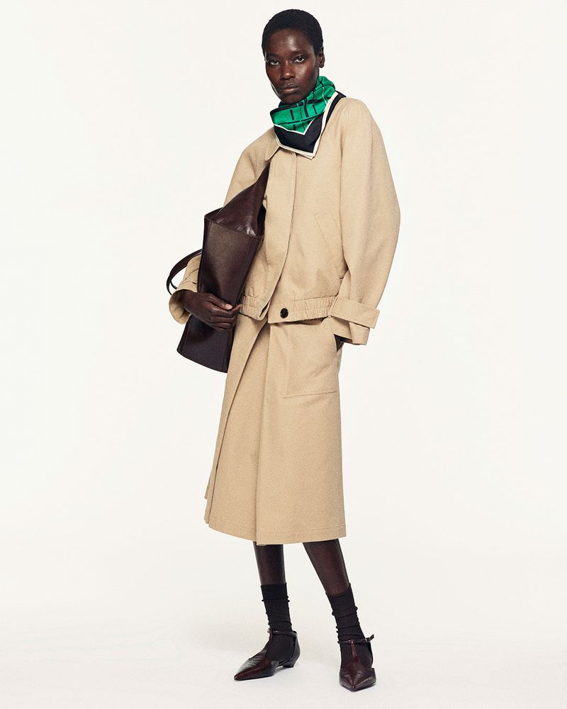 Fashion editorial shot of an African-descent adult woman in a beige skirt and jacket, holding a brown tote bag against a plain white backdrop.
