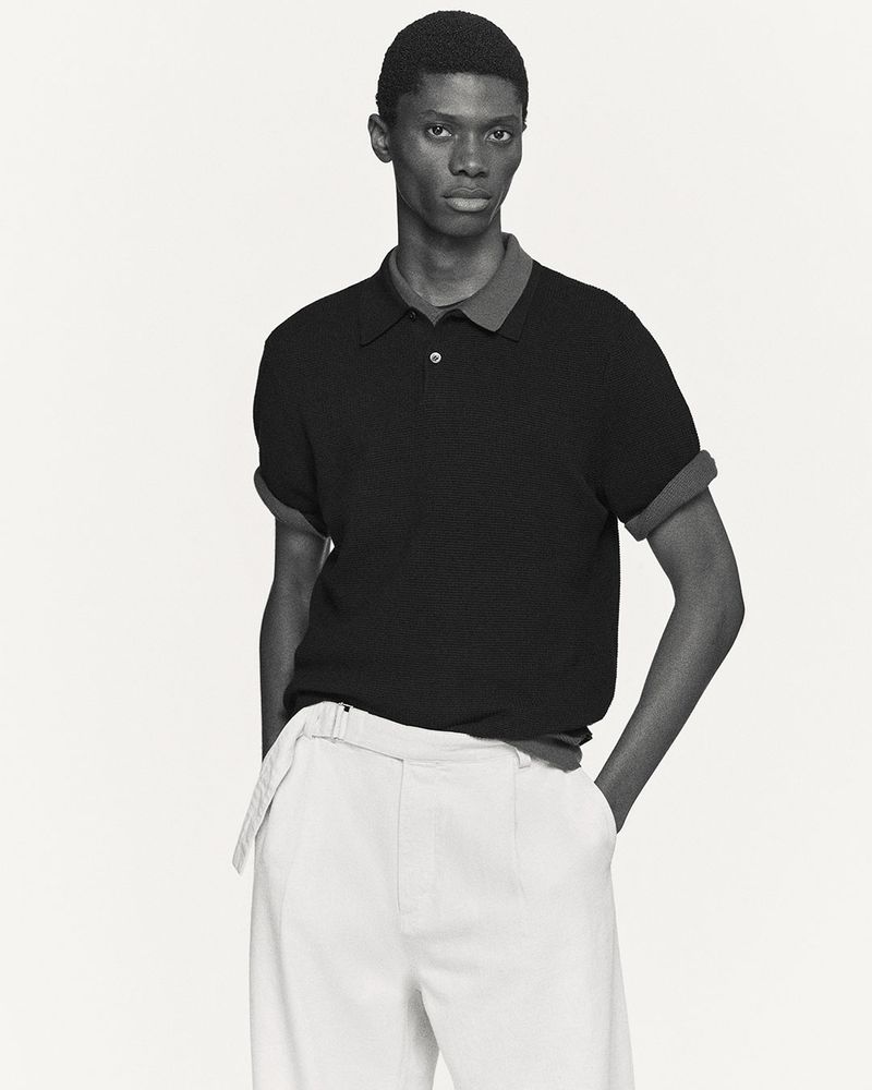 Monochrome fashion shot: a dark-skinned male model poses in a black polo shirt and white trousers against a bright, neutral backdrop.