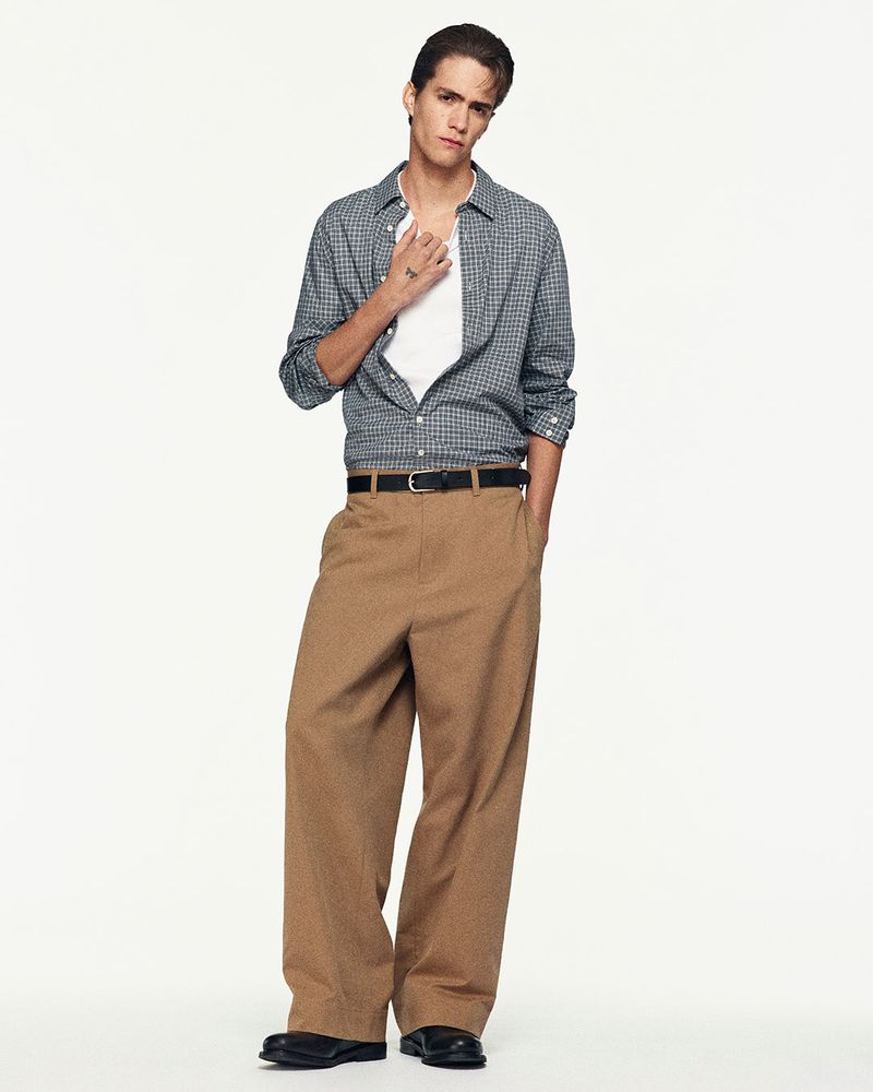 Full-length shot of a young adult male model in a checked shirt and khaki pants, standing against a plain white studio backdrop.