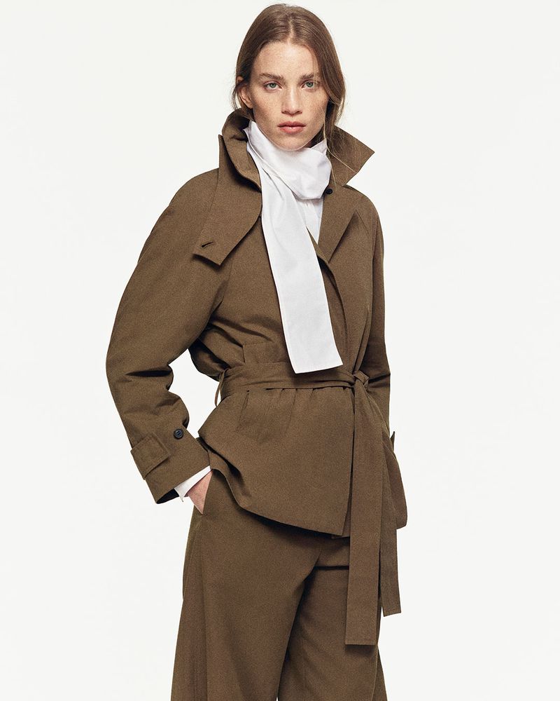 Fashion model in a brown trench coat and matching pants poses against a white backdrop in a studio setting for a fashion editorial.