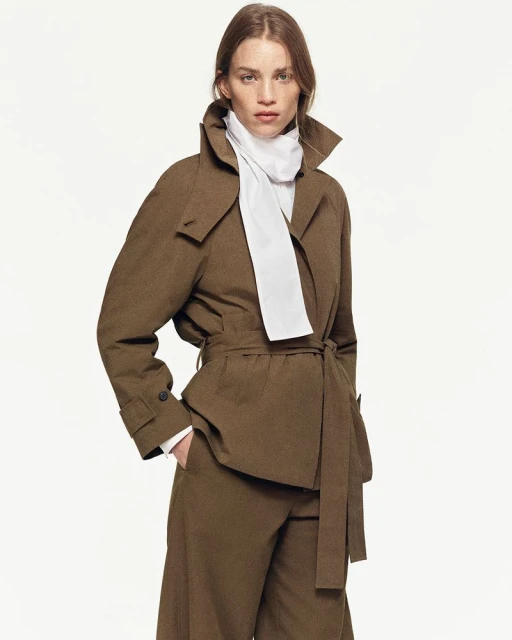 Fashion model in a brown trench coat and matching pants poses against a white backdrop in a studio setting for a fashion editorial.