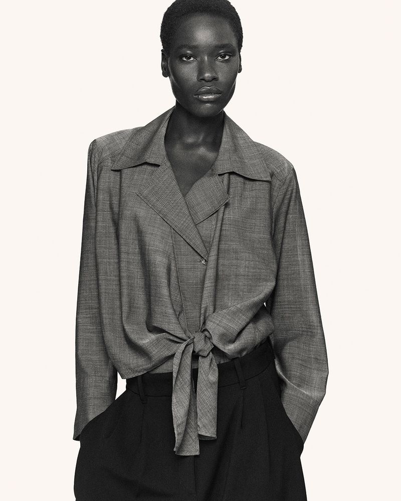 Monochrome fashion shot of a Black woman wearing a grey tie-front shirt and black slacks, posed against a neutral backdrop.
