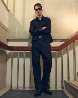 Model wearing a dark jacket and trousers with sunglasses stands on a staircase with a wooden handrail for the COS Spring 2026 lookbook.