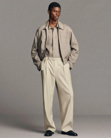 Black man wearing a beige jacket, matching shirt, and light trousers standing against a solid grey studio background.